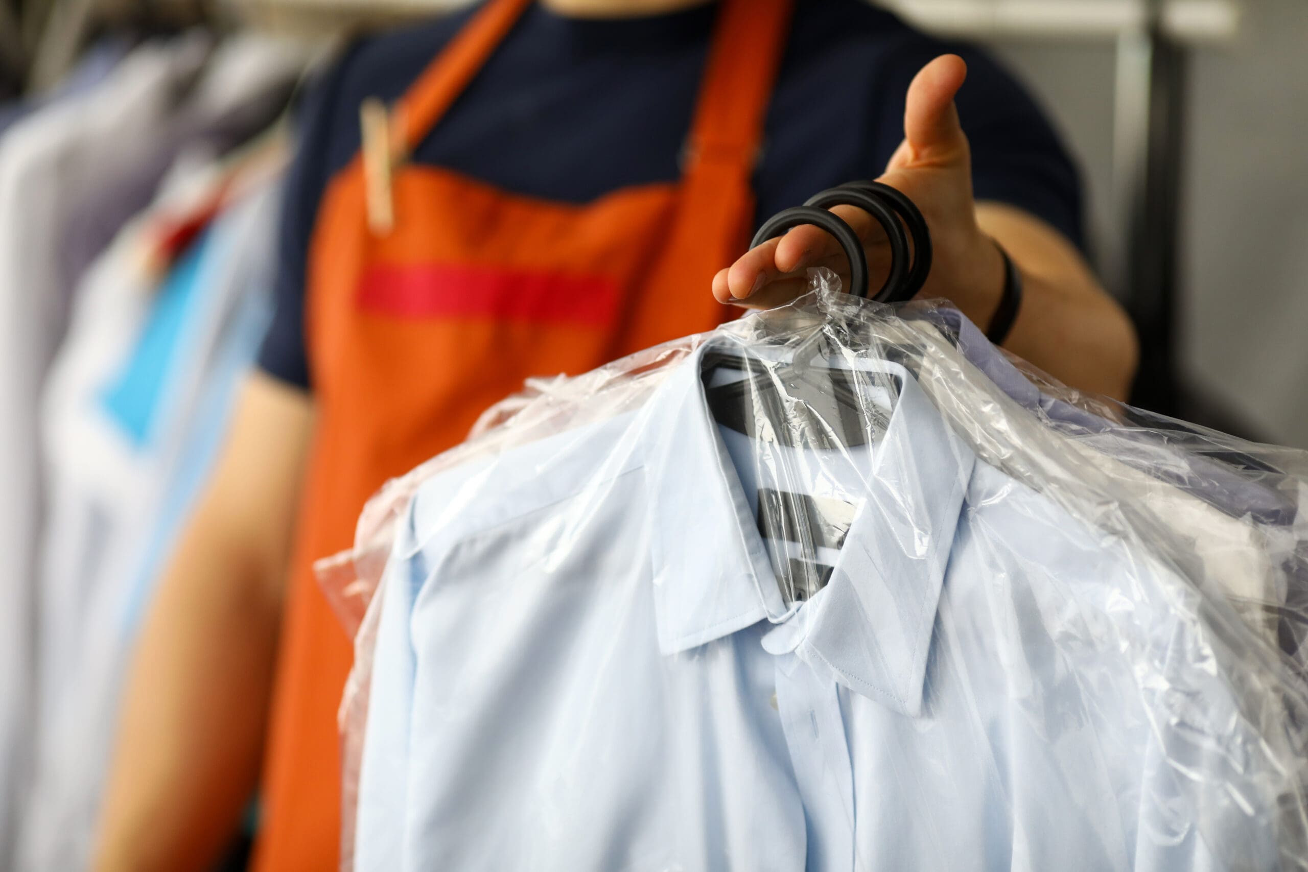 A person wearing an orange apron holds out a dry-cleaned blue dress shirt covered in a plastic garment bag. The focus is on the shirt, with more clothing in plastic in the blurred background.