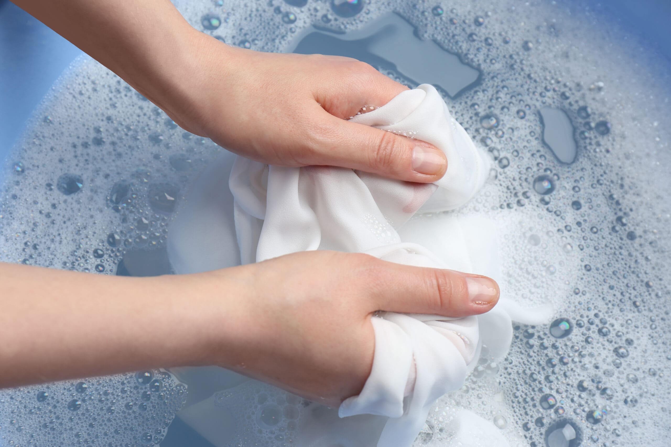 Hands washing a white cloth in soapy water, with bubbles forming on the surface. The scene suggests a gentle, manual laundry process.