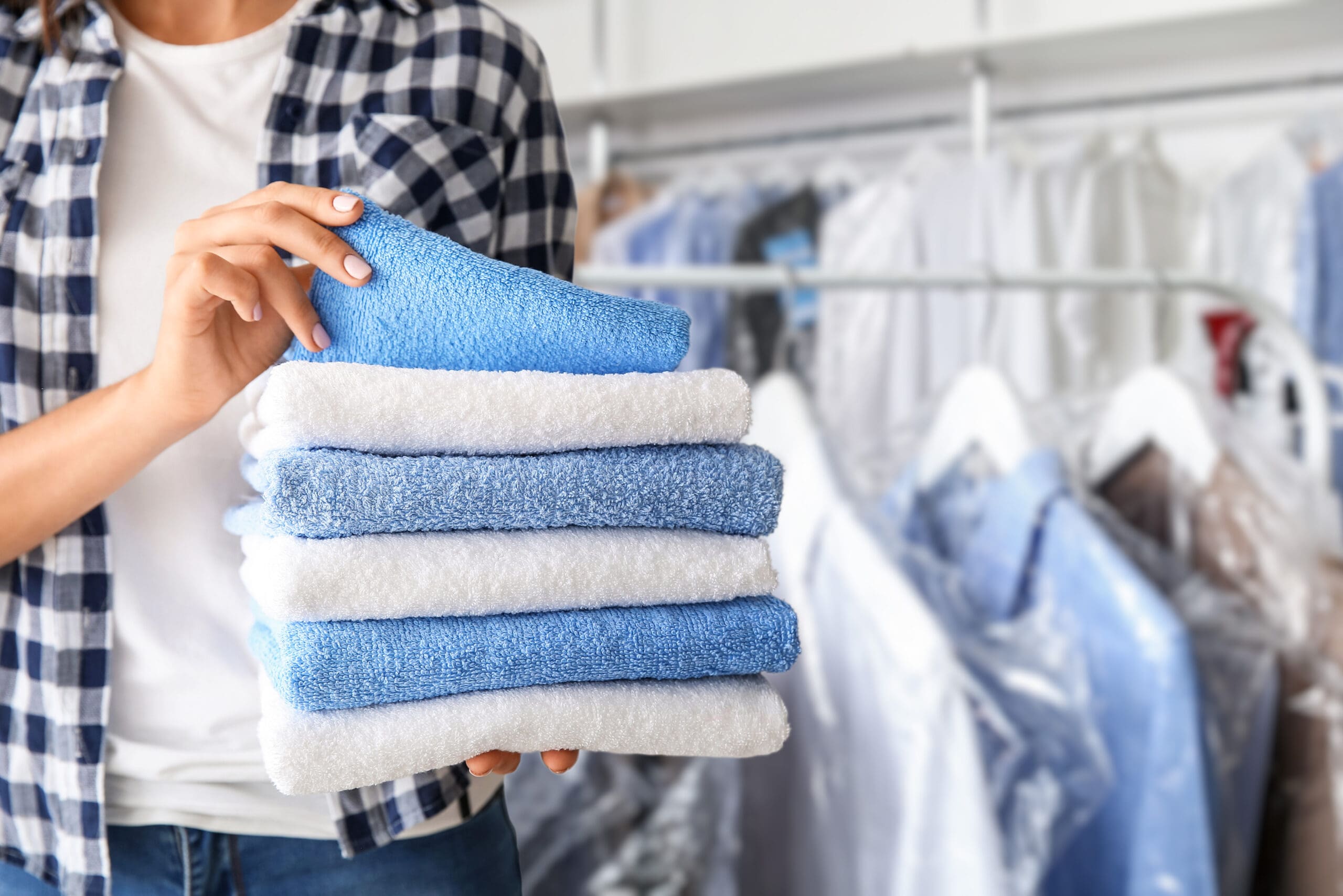 A person in a plaid shirt holds a stack of folded blue and white towels in a laundry room. Racks of hanging clothes are visible in the background.