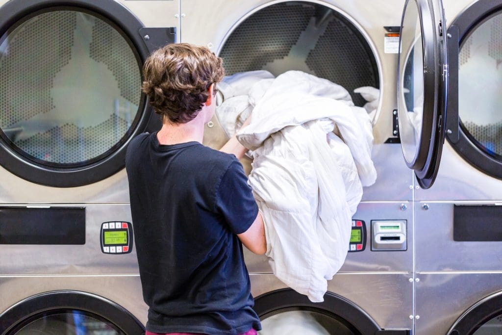 A person in a black shirt loads a large white comforter into a front-loading dryer at a laundromat. The dryer has digital displays and is part of a row of similar machines.