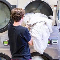 A person in a black shirt loads a large white comforter into a front-loading dryer at a laundromat. The dryer has digital displays and is part of a row of similar machines.