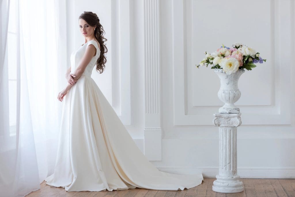 A woman in an elegant white wedding dress stands near a sunlit window in a bright room with white paneling. Her dress has a long train flowing on the floor. A decorative white urn with colorful flowers is placed on a pedestal nearby.