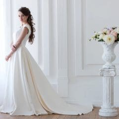 A woman in an elegant white wedding dress stands near a sunlit window in a bright room with white paneling. Her dress has a long train flowing on the floor. A decorative white urn with colorful flowers is placed on a pedestal nearby.