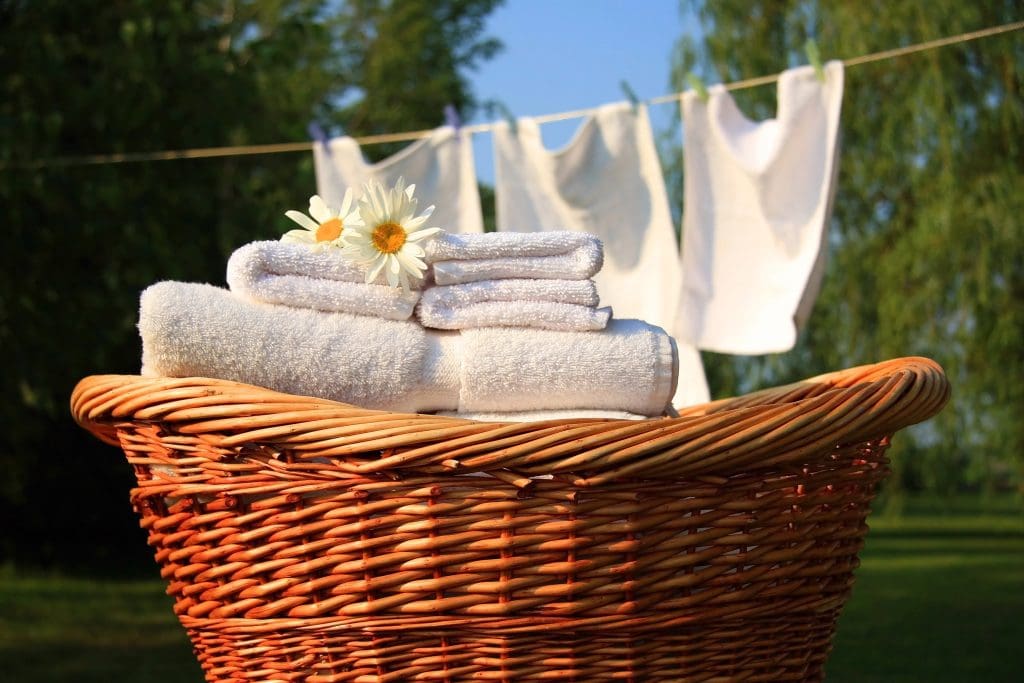 A wicker basket filled with neatly folded white towels, adorned with daisies, is in the foreground. In the background, three white cloths hang on a clothesline against a sunny, green outdoor setting.