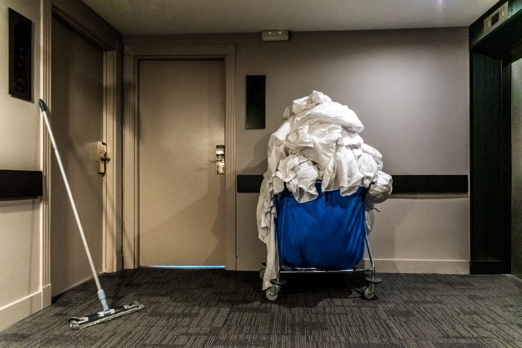 A hallway with a mop leaning against a wall, a closed door, and a blue cart overflowing with white linens. The walls are beige, and the floor is carpeted in gray.