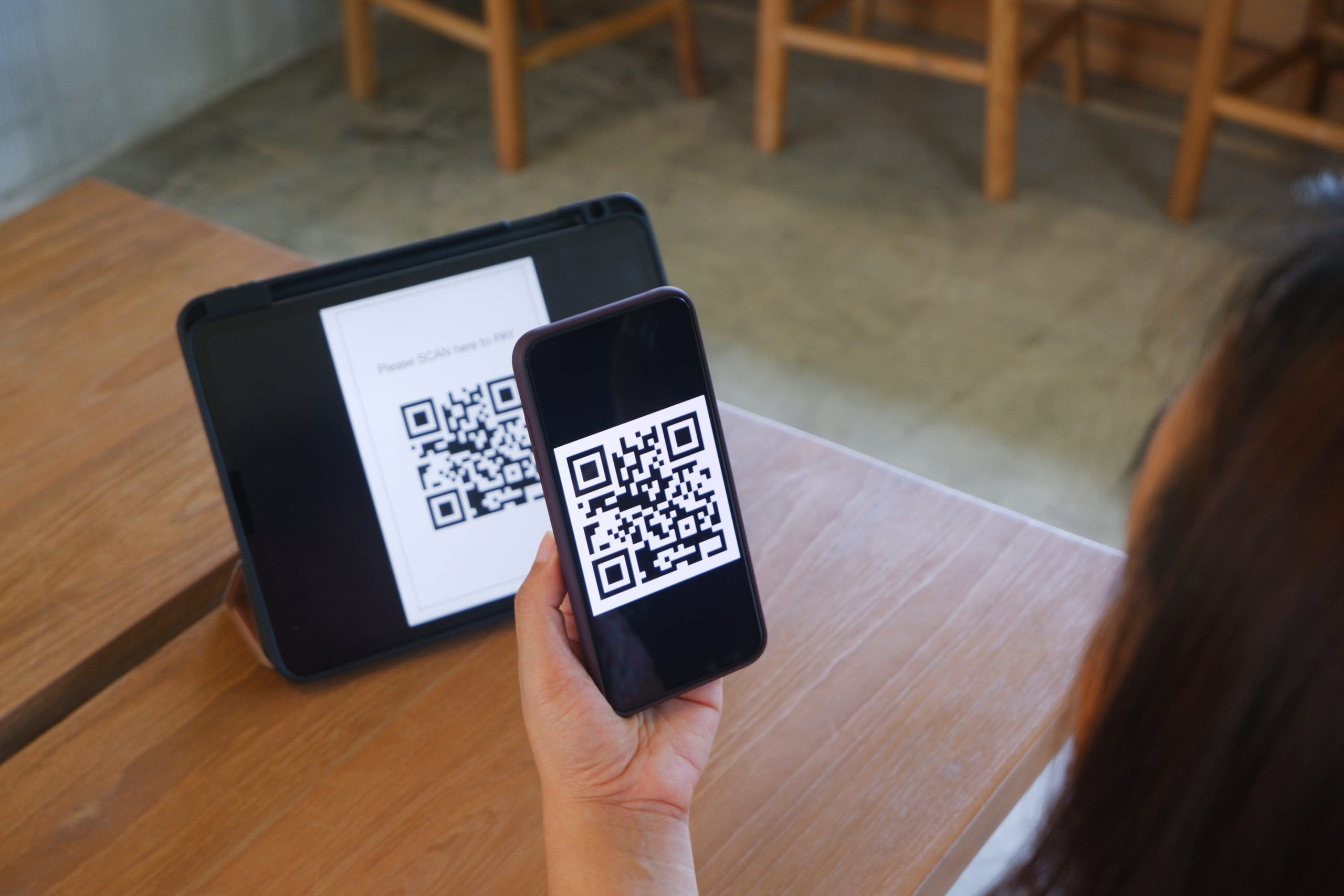 A person scans a QR code displayed on a tablet using a smartphone in a cafe setting. Wooden chairs and a table made of light wood are visible in the background.