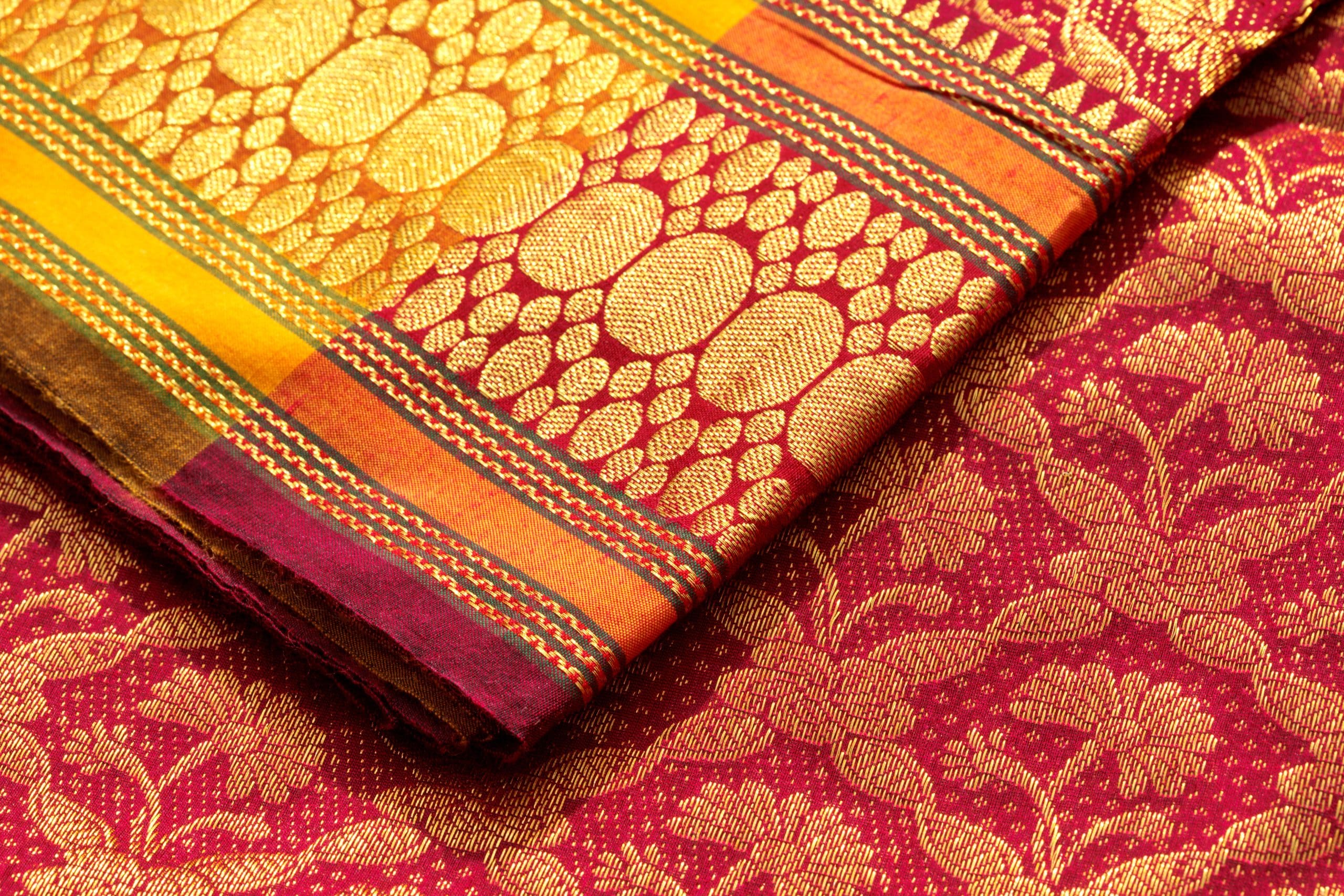 Close-up of a vibrant, intricately woven Indian silk sari. The fabric features golden floral and geometric patterns on a rich maroon base, with colorful striped borders in yellow, pink, and green hues.