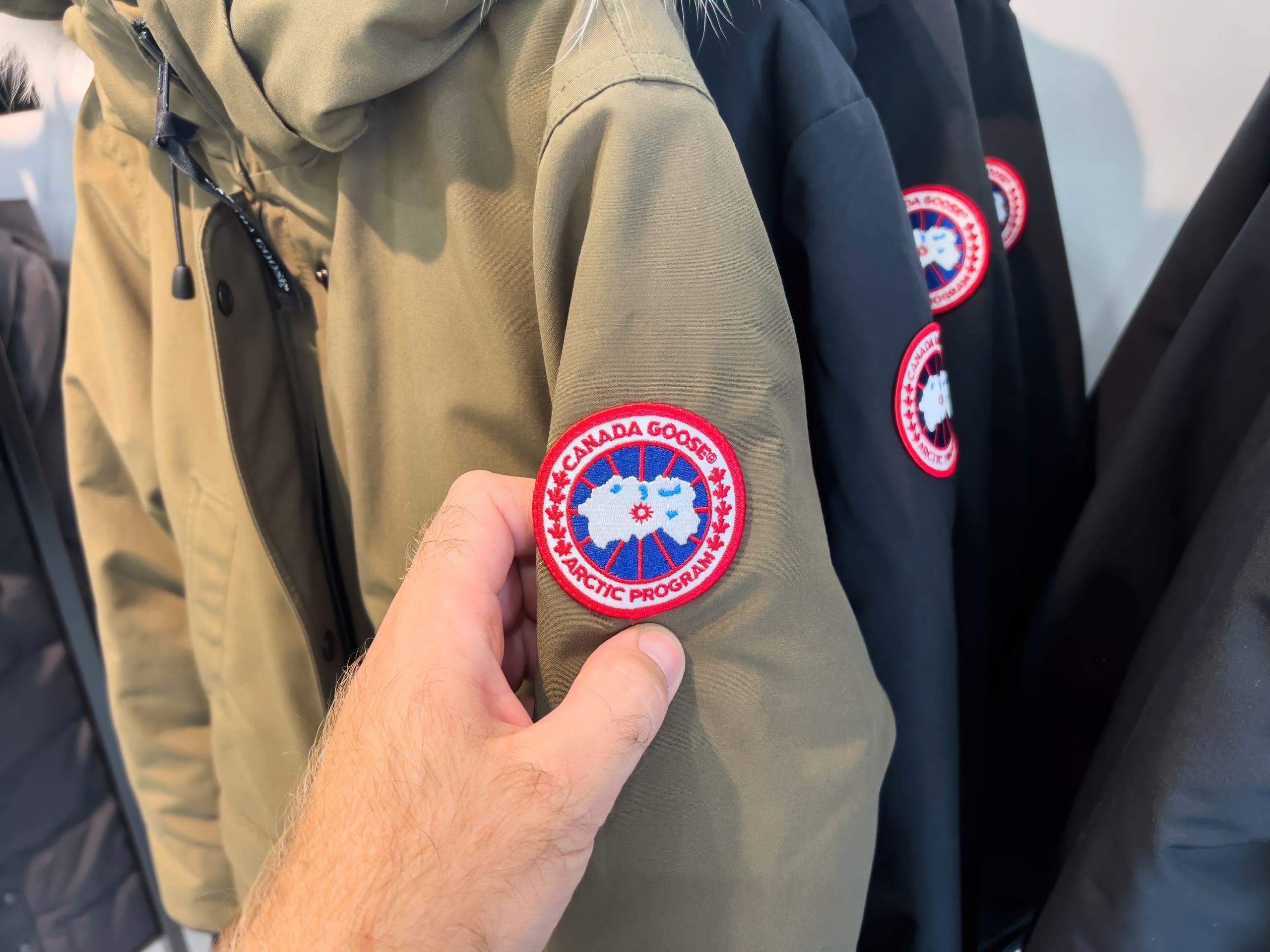 A person is holding the sleeve of a beige jacket featuring a circular patch with the words "Canada Goose Arctic Program" and a red and blue emblem. Other jackets hang in the background.