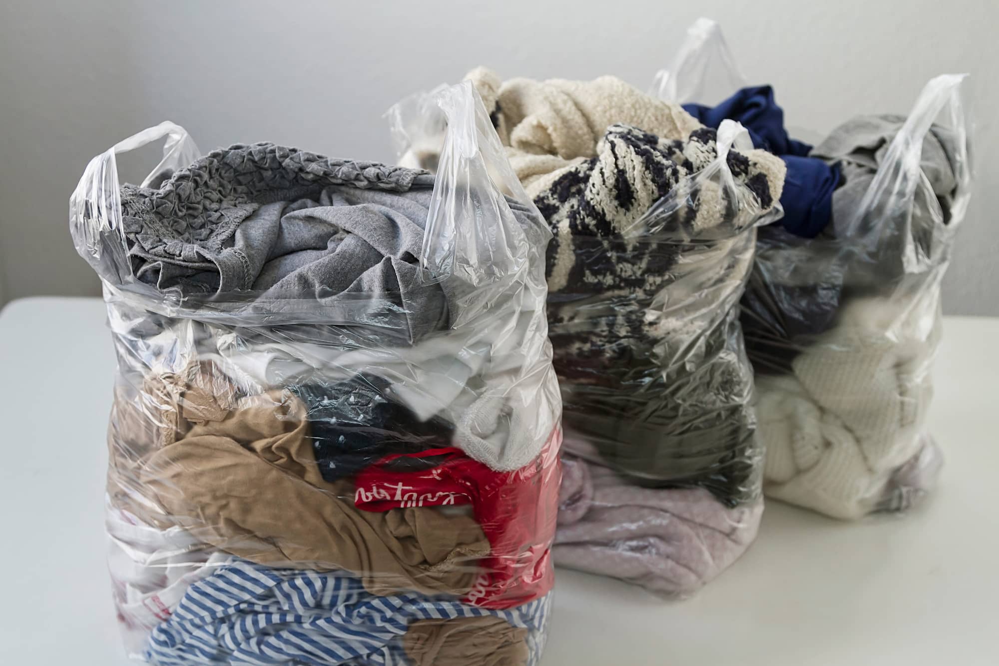 Three clear plastic bags filled with assorted folded clothes, including items in gray, beige, blue, red, and striped patterns, are placed on a white surface against a plain background.