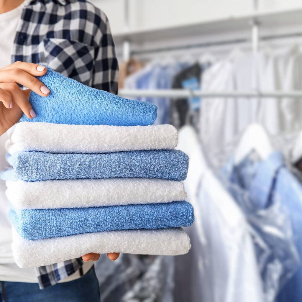 A person in a plaid shirt holds a neatly folded stack of blue and white towels in a laundry or dry cleaning setting, with hanging clothes visible in the background.