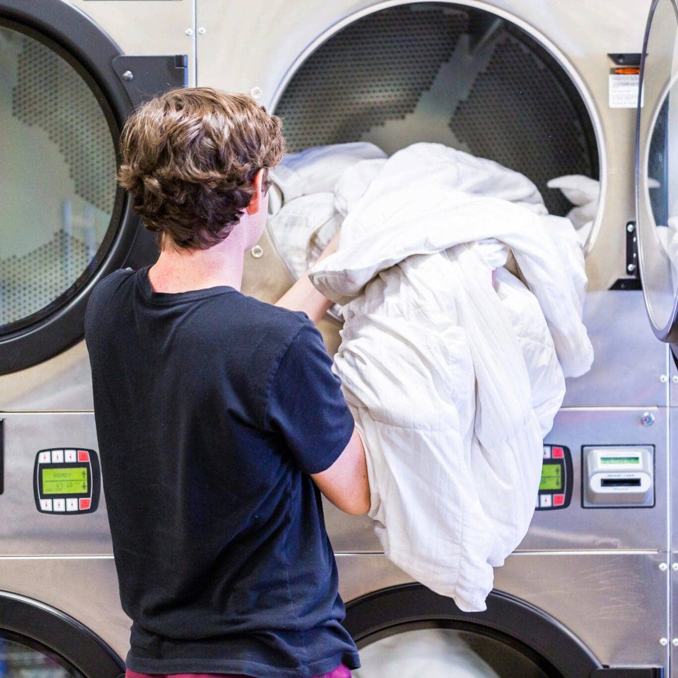 A person with short brown hair, wearing a black shirt, loads a large white comforter into a front-loading industrial dryer at a laundromat.