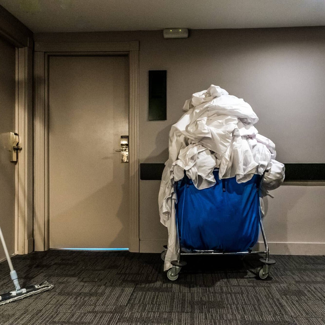 A large blue laundry cart overflowing with white linens sits in a hallway next to a closed beige door; a mop is partially visible on the left side of the image.
